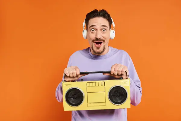 Sorpreso uomo barbuto in maglione viola e cuffie wireless che tengono boombox su sfondo arancione — Foto stock