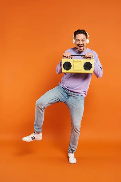 Cheerful bearded man in purple sweater and wireless headphones holding boombox on orange background — Stock Photo