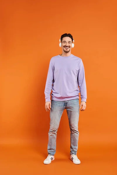 Cheerful bearded man in purple sweater and wireless headphones listening music on orange background — Stock Photo