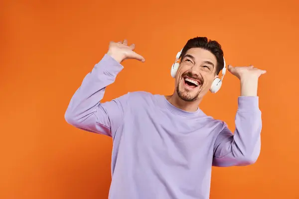 Thrilled bearded man in purple sweater and wireless headphones listening music on orange background — Stock Photo