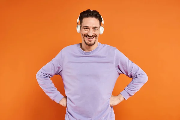 Homem alegre em camisola roxa e fones de ouvido sem fio ouvir música no fundo laranja — Fotografia de Stock