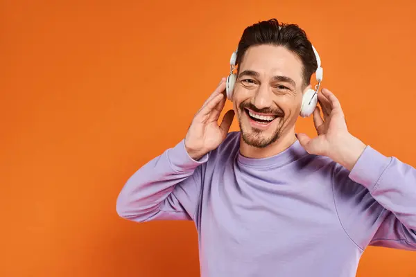 Hombre positivo en suéter morado y auriculares inalámbricos escuchando música sobre fondo naranja - foto de stock