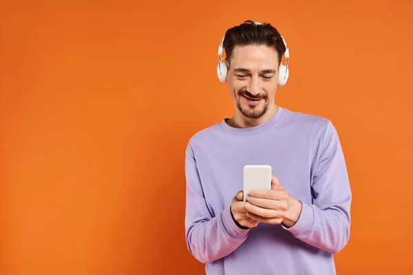 Hombre positivo en auriculares inalámbricos escuchando música y utilizando el teléfono inteligente sobre fondo naranja - foto de stock