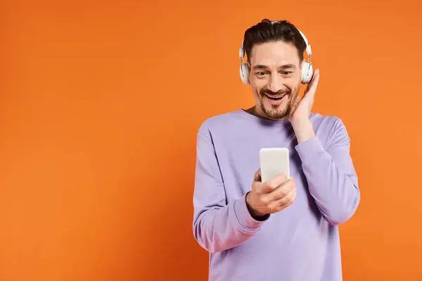 Hombre sonriente en auriculares inalámbricos escuchando música y usando un teléfono inteligente sobre fondo naranja - foto de stock