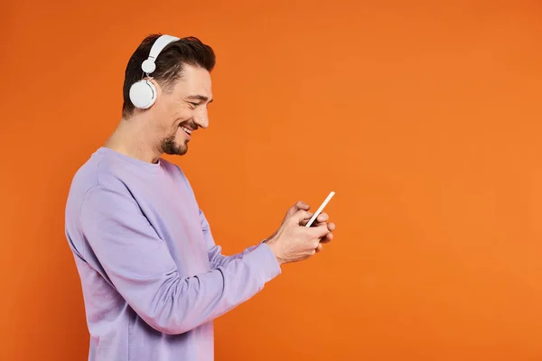 Hombre feliz en auriculares inalámbricos escuchando música y mensajes de texto en el teléfono inteligente sobre fondo naranja - foto de stock