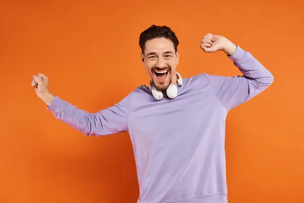 Joyful man in purple sweater with wireless headphones posing on orange background, music lover — Stock Photo