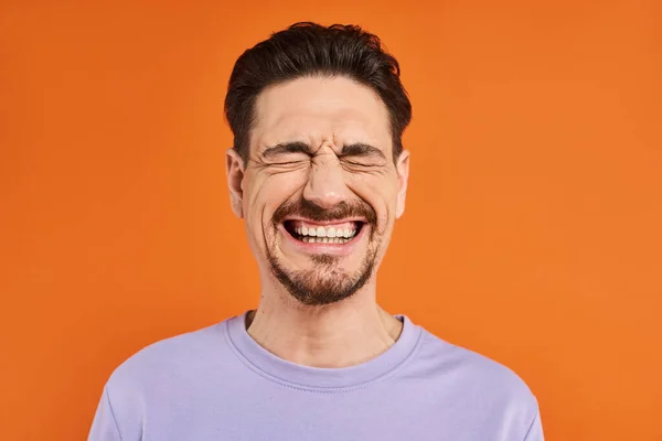 Счастье, взволнованный бородатый мужчина в фиолетовой толстовке улыбается с закрытыми глазами на оранжевом фоне — стоковое фото