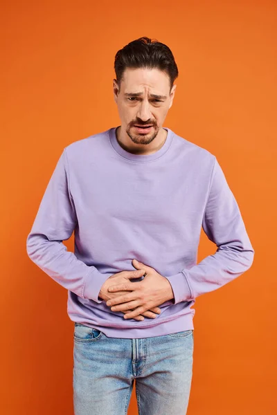 Homme barbu en pull violet et jeans souffrant de maux d'estomac sur fond orange, douleur — Photo de stock