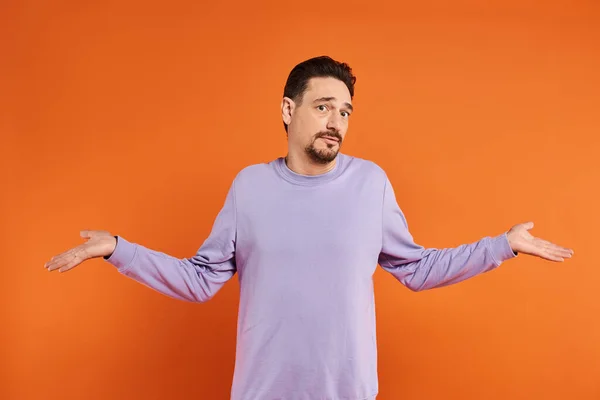 Hombre barbudo confuso en suéter púrpura mostrando gesto encogiéndose de hombros con las manos sobre fondo naranja - foto de stock