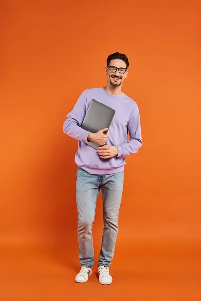 Longitud completa del hombre barbudo en gafas y suéter púrpura celebración portátil sobre fondo naranja - foto de stock
