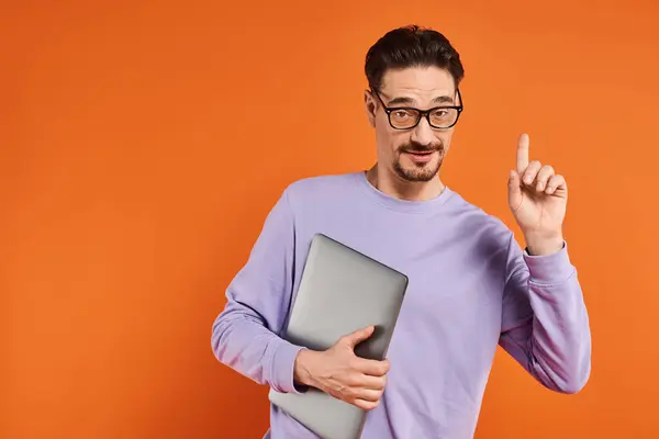 Homme barbu dans des lunettes et pull violet tenant ordinateur portable et pointant vers le haut sur fond orange — Photo de stock