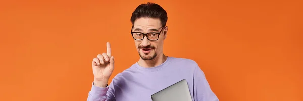 Homme barbu dans des lunettes et pull violet tenant ordinateur portable et pointant vers le haut sur fond orange, bannière — Photo de stock