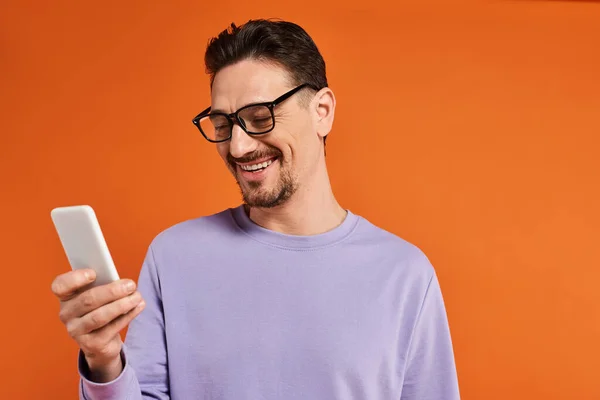 Uomo allegro in occhiali e maglione viola utilizzando smartphone su sfondo arancione, sms — Foto stock
