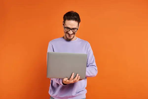 Homme barbu en lunettes et pull violet à l'aide d'un ordinateur portable sur fond orange, travail à distance — Photo de stock