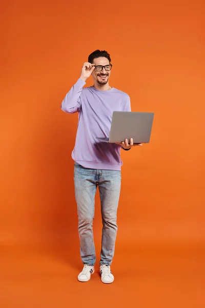 Веселый человек в очках и фиолетовый свитер с помощью ноутбука на оранжевом фоне, удаленная работа — стоковое фото