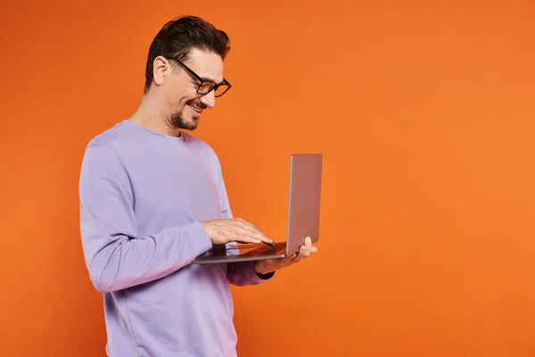 Улыбающийся мужчина в очках и фиолетовом свитере с помощью ноутбука на оранжевом фоне, удаленная работа — стоковое фото
