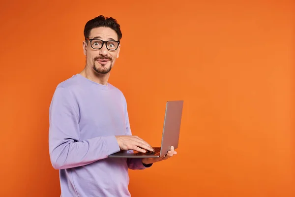 Здивований чоловік в окулярах і фіолетовий светр, використовуючи ноутбук на помаранчевому фоні, віддалена робота — стокове фото