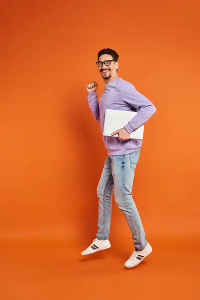 Homme barbu heureux dans les lunettes et le pull violet marche avec ordinateur portable sur fond orange — Photo de stock