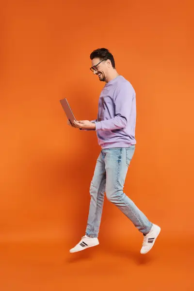 Homme joyeux dans les lunettes et le pull violet en utilisant un ordinateur portable sur fond orange, travail à distance — Photo de stock