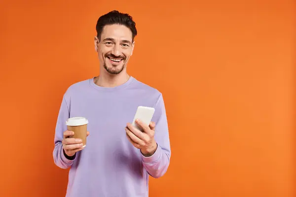 Homme souriant dans des lunettes tenant du café à emporter et utilisant un smartphone sur fond orange, caféine — Photo de stock