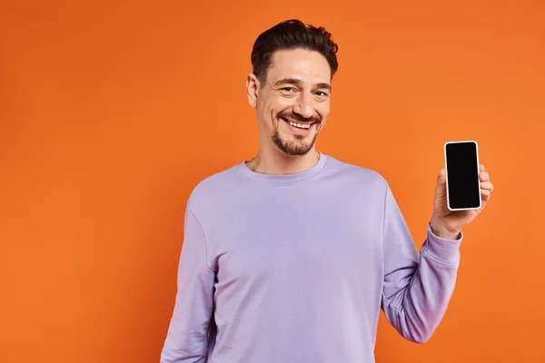 Щасливий чоловік в окулярах і фіолетовий светр тримає смартфон з порожнім екраном на помаранчевому фоні — стокове фото