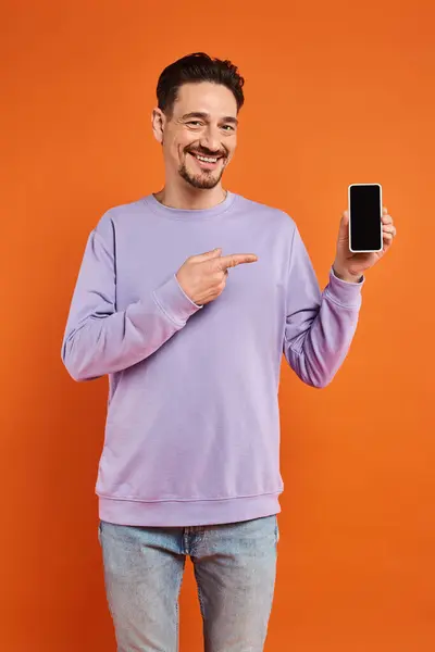 Hombre feliz en gafas y suéter púrpura apuntando al teléfono inteligente con pantalla en blanco sobre fondo naranja - foto de stock
