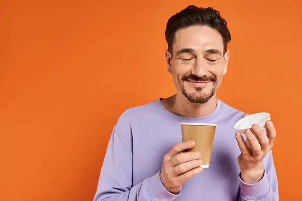 Homme gai avec barbe tenant tasse en papier avec café pour aller sur fond orange, plaisir — Photo de stock