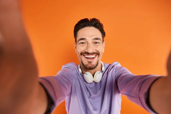 Homem barbudo feliz em fones de ouvido sem fio ouvindo música e olhando para a câmera no fundo laranja — Fotografia de Stock