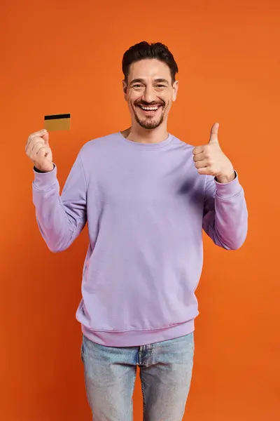 Homme gai avec barbe poussant pouce vers le haut et tenant la carte de crédit sur fond orange, comme — Photo de stock