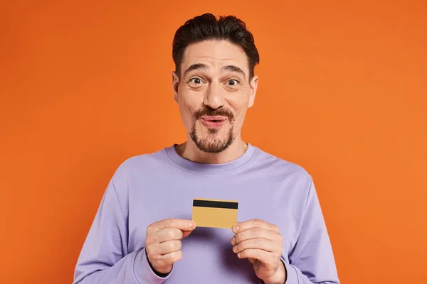 Homme gai avec barbe souriant et tenant la carte de crédit sur fond orange, regardant la caméra — Photo de stock