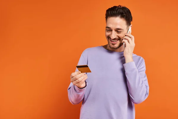 Homme joyeux souriant et tenant carte de crédit tout en parlant sur smartphone sur fond orange — Photo de stock