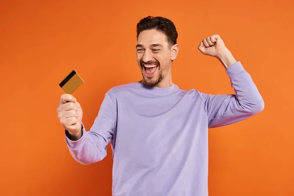 Homme excité avec la barbe souriant et tenant la carte de crédit sur fond orange, geste réjouissant — Photo de stock