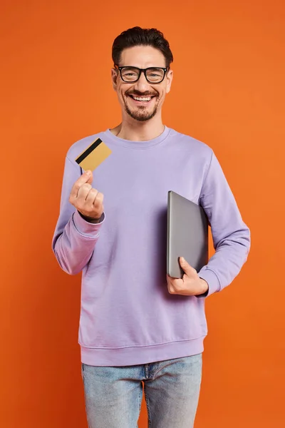 Homme gai dans des lunettes debout avec carte de crédit et ordinateur portable sur fond orange, achats en ligne — Photo de stock