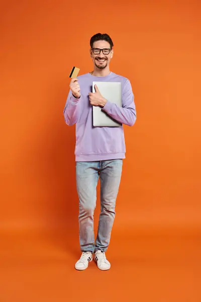 Homme heureux dans des lunettes debout avec carte de crédit et ordinateur portable sur fond orange, achats en ligne — Photo de stock