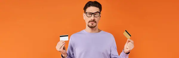 Hombre escéptico en gafas y suéter púrpura comparando dos tarjetas de crédito sobre fondo naranja, bandera - foto de stock