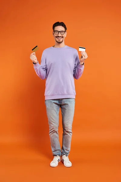 Homem barbudo feliz em óculos segurando cartões de crédito no fundo laranja, compras e consumismo — Fotografia de Stock