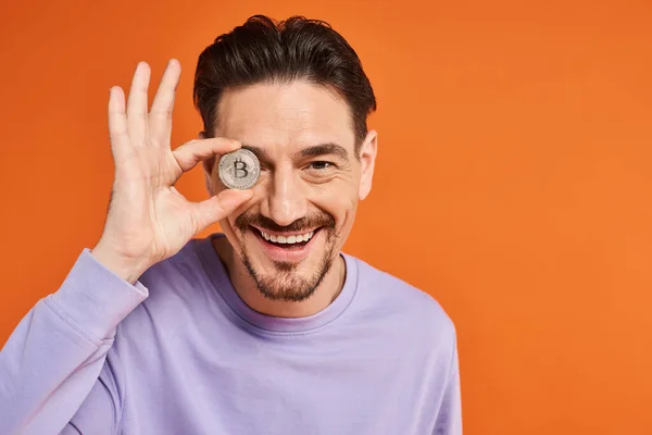 Uomo felice tenendo bitcoin sopra l'occhio e sorridendo alla fotocamera su sfondo arancione, criptovaluta — Foto stock