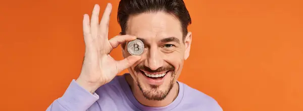 Homme heureux tenant Bitcoin près de l'œil et souriant à la caméra sur fond orange, bannière crypto-monnaie — Photo de stock