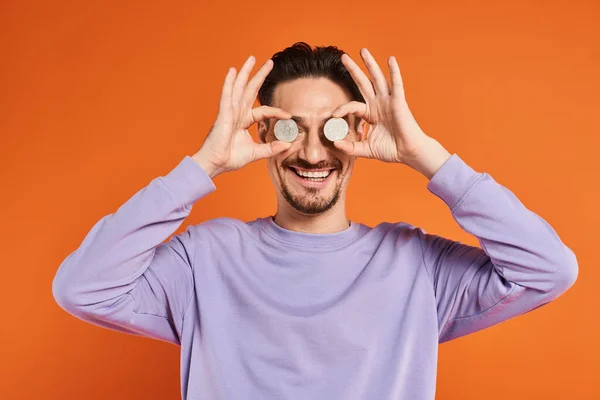 Счастливый человек, держащий биткоины над глазами и улыбающийся камере на оранжевом фоне, криптовалюта — стоковое фото