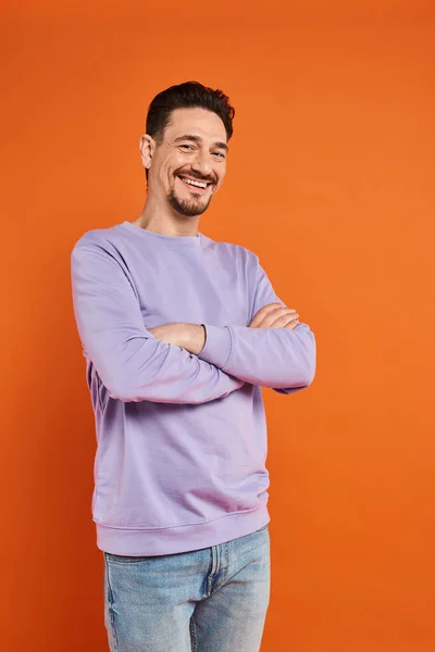 Happy man in purple sweater and jeans standing with crossed arms on orange background, casual wear — Stock Photo
