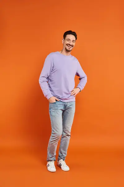 Felice uomo in maglione viola e jeans in piedi con mano in tasca su sfondo arancione, abbigliamento casual — Foto stock