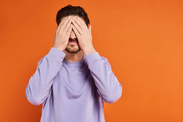 Homem com barba em camisola casual roxo escondendo o rosto com as mãos no fundo laranja, humor brincalhão — Fotografia de Stock