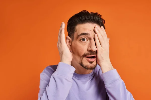 Hombre sorprendido con barba en suéter púrpura ocultar la cara con la mano sobre fondo naranja, humor juguetón - foto de stock