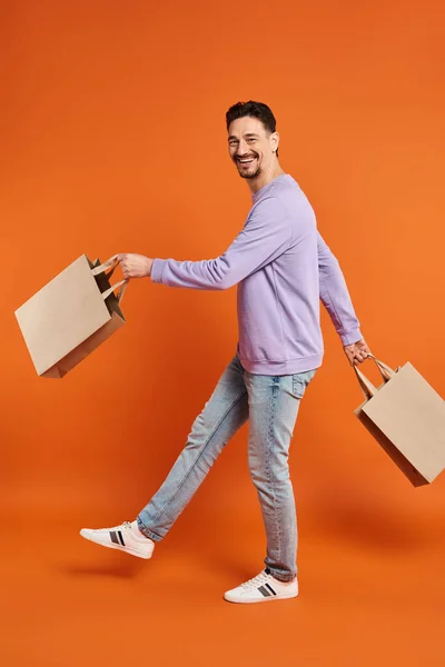 Comprimento total de homem barbudo feliz em traje casual andando com sacos de compras no fundo laranja — Fotografia de Stock