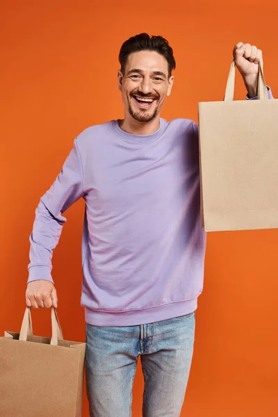 Cheerful bearded man in casual attire holding shopping bags on orange background, consumerism — Stock Photo