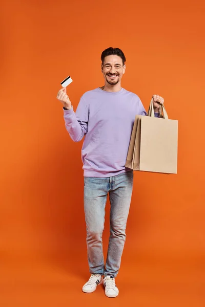 Hombre barbudo alegre en traje casual sosteniendo bolsas de compras y tarjeta de crédito sobre fondo naranja - foto de stock