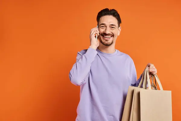 Smiling man talking on the phone and carrying shopping bags on orange background, consumer — Stock Photo