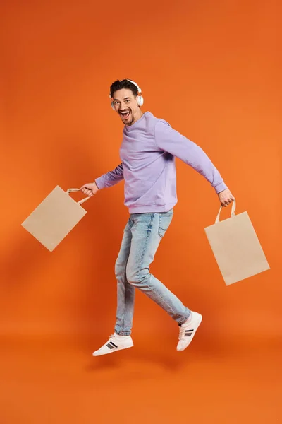 Joyful man in wireless headphones levitating with shopping bags on orange background, purchase — Stock Photo