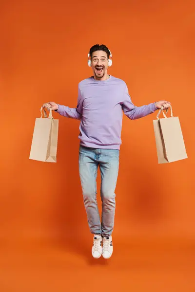 Excited man in wireless headphones levitating with shopping bags on orange background, purchase — Stock Photo