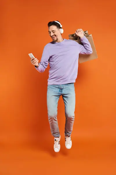 Hombre excitado en auriculares levitando con bolsas de compras y sosteniendo el teléfono inteligente sobre fondo naranja - foto de stock